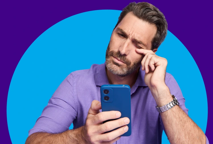 Man in a blue squinting while looking at his phone and rubbing his eye with his index finger